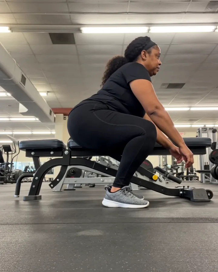 A woman at the bottom of a bodyweight squat beside a bench in an ordinary gym, filmed from the floor