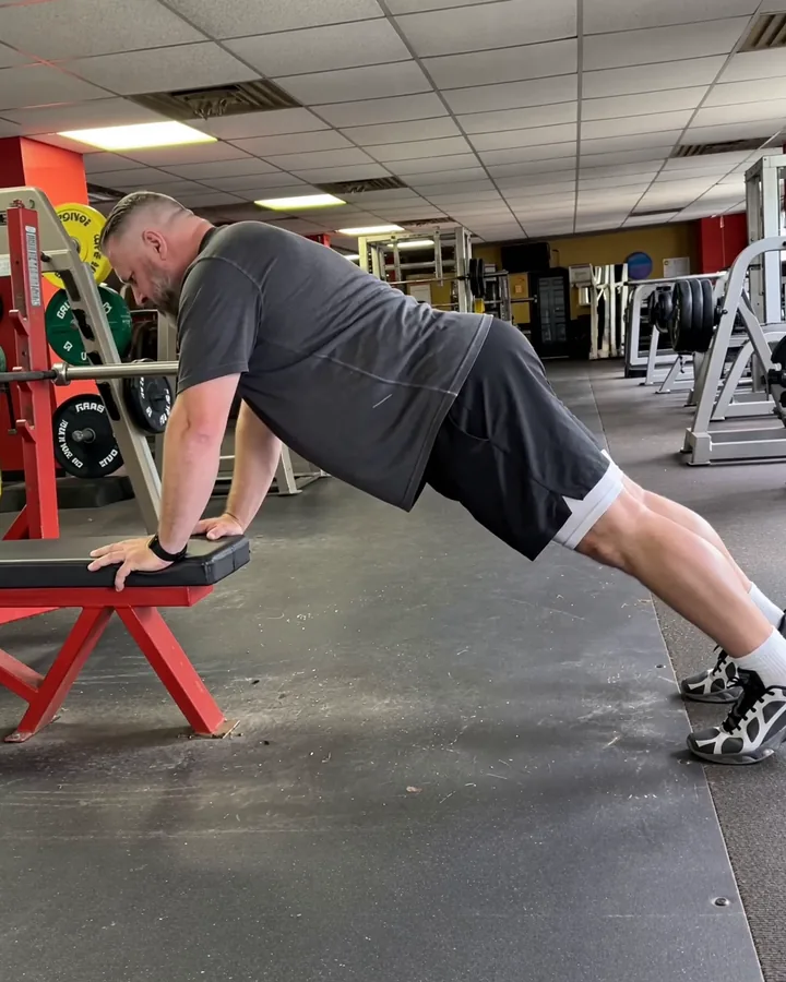 A man doing incline push-ups with his hands on a gym bench, side-on, whole body in frame